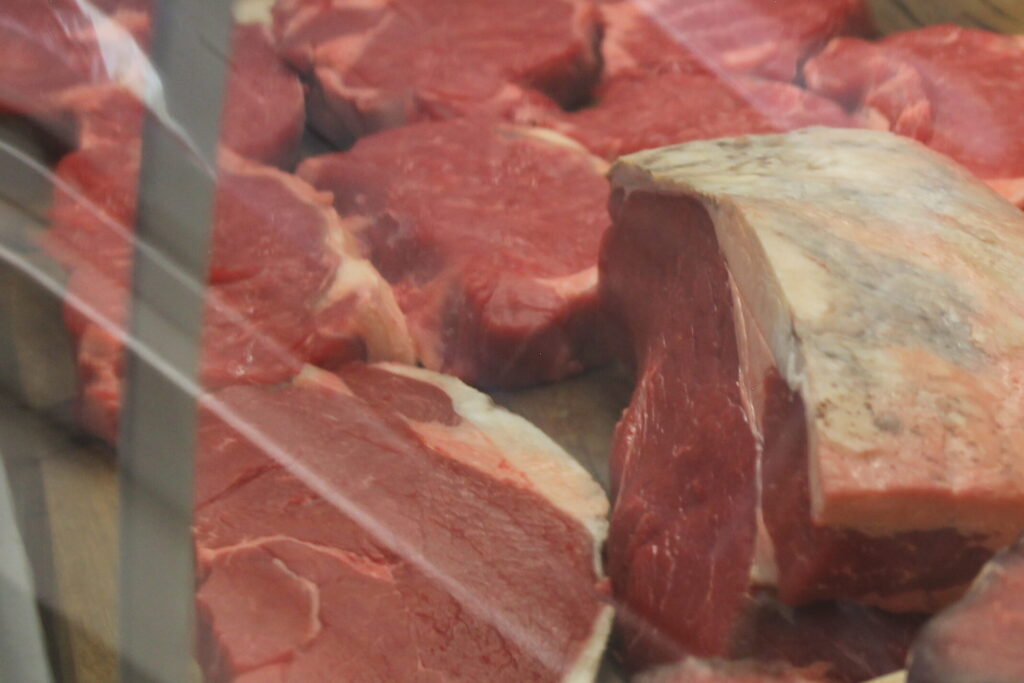 This image displays a selection of raw meat cuts in a refrigerated display case, likely in a butcher shop or gourmet grocery store. The meat includes various types and cuts, such as thick steaks and large roasts, with visible marbling and fat caps. The cuts are arranged neatly on paper with a logo, giving a clean, professional presentation. The glass display case reflects some light, adding to the freshness and quality-focused atmosphere of the store. The meats appear to be high-quality, with rich red colors, suggesting they’re fresh and possibly sourced from premium suppliers.
